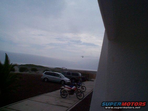 Two dirt bikes parked on a paved surface with a van and SUV in the background near a beach