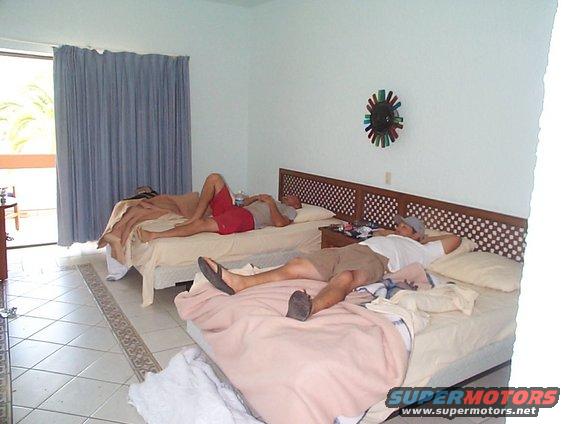 Two guys are passed out on hotel beds, one in red shorts, the other in a white shirt and tan shorts