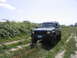 2000 Jeep Cherokee - A dark green Jeep Cherokee drives on a dirt path surrounded by vegetation under a partly cloudy sky.