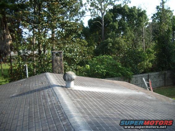 Roof with asphalt shingles, a brick chimney, and a metal turbine vent. Trees and a wooden fence are in the background