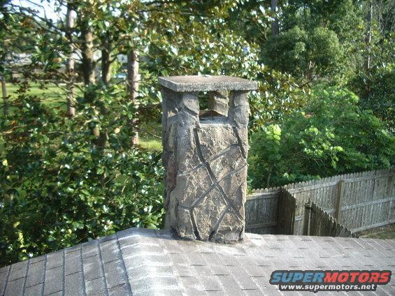 Stone chimney on asphalt shingle roof with wooden fence and trees in background