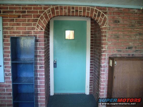 Brick archway over a teal door with a small window. A metal shelf is to the left, and a brown cabinet is to the right