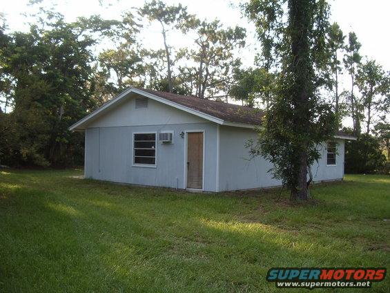 small light blue house with a window air conditioner and a wooden door, surrounded by grass and trees