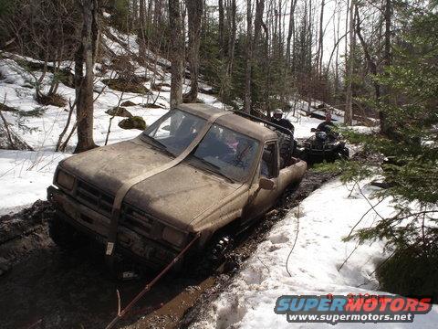 Mud-covered pickup truck stuck in a muddy trail with snow on the sides, an ATV behind it