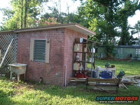 Backyard scene with a brick shed, louvered window, outdoor sink, and metal shelves holding various containers. A wooden bench sits in front of the shed