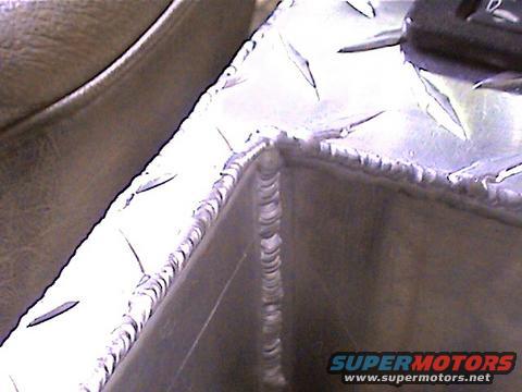 Close-up of a diamond plate aluminum corner with visible welds. Supermotors.net logo in lower right