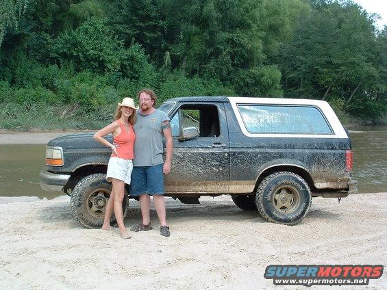 Couple posing by a muddy 1993 Ford Bronco on a sandy riverbank