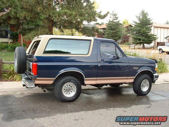 Blue and tan Ford Bronco parked on a wet street, spare tire on the back