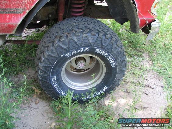 Close-up of a large off-road tire on a vehicle, showing suspension components and part of the red body. Tire reads 'BAJA BELTED MICKEY THOMPSON'