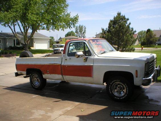 White and orange pickup truck parked in driveway, for sale sign in window. Another white truck is visible in the background