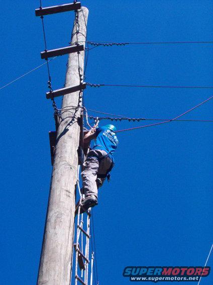 Lineman in blue shirt and grey pants climbing a wooden utility pole with a rope ladder, blue sky