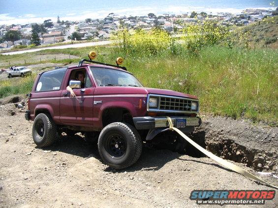 Maroon 1985 Ford Bronco II with roof rack and off-road tires stuck in dirt, being pulled by a strap. White pickup truck in background