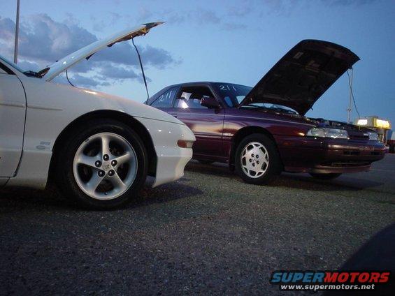 White car on left, maroon car on right, both with hoods open. Sky is blue with clouds