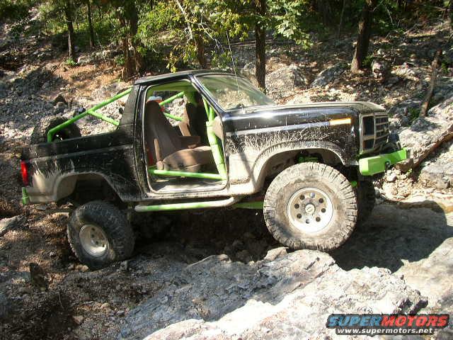 Black off-road truck with green roll cage and lime green bumper, seen from the front-right on rocky terrain