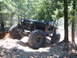 1988 Ford Bronco - A heavily modified black Jeep Wrangler navigates a rocky, wooded trail with its large tires.