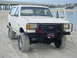 1989 Ford Bronco - A white 1989 Ford Bronco with off-road tires parked on a sandy beach, with another vehicle visible in the background.