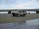 1989 Ford Bronco - A classic Ford Bronco sits on the sand near the water, with a bridge overhead and another vehicle in the distance.