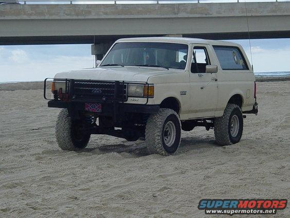 White Ford Bronco SUV with a black bull bar and off-road tires on a sandy beach