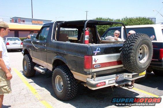 Black Ford Bronco with lifted suspension and spare tire on back, parked next to another SUV. Hooters in background