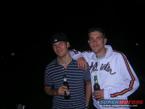 Two men stand together outdoors at night, each holding a beer bottle