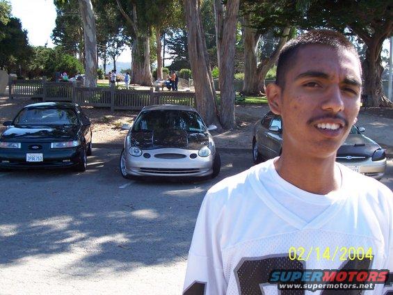 guy in front of two cars, one is a silver Ford Taurus with a black hood, the other is a dark green sedan