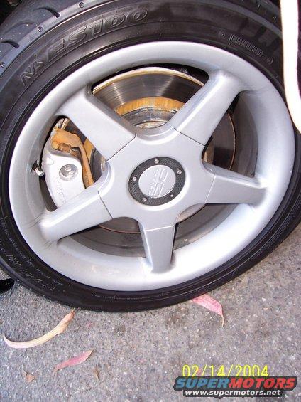 Silver five-spoke wheel with a brake caliper and rotor visible, on a tire with text