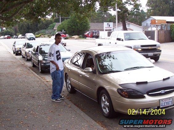 Gold Ford Taurus parked on street, guy in Raiders jersey standing next to it. Other cars lined up behind