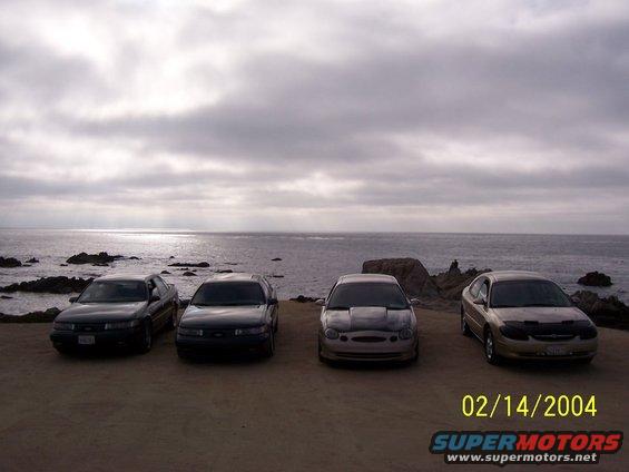 Four sedans parked on a dirt lot overlooking the ocean, one is a 1998 Mercury Sable