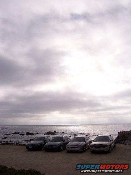 four sedans parked on dirt lot facing ocean, cloudy sky