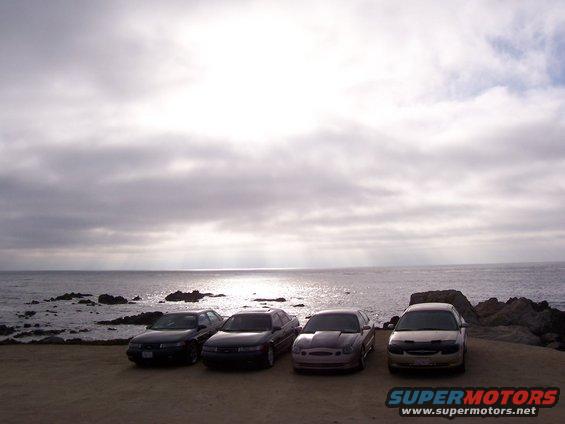 Four sedans parked on a dirt lot by the ocean, with a cloudy sky