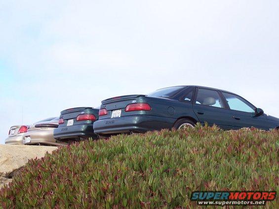 Three sedans parked on a grassy hill, two are dark green, one is tan. The rear of the vehicles are visible