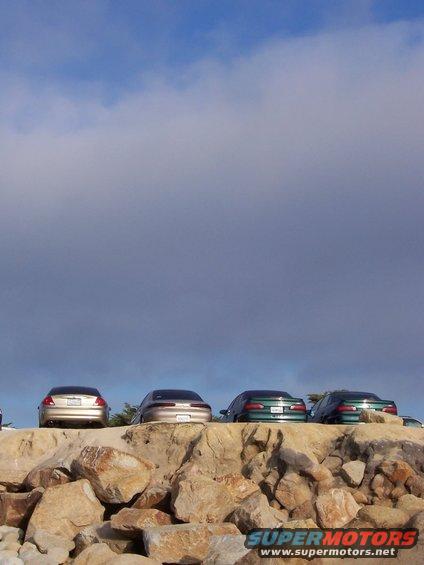 four sedans parked on a rocky ledge under a cloudy sky