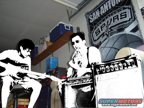 Two guys are playing guitars in a garage. One is sitting and playing a guitar. The other is standing and playing a guitar next to an amplifier