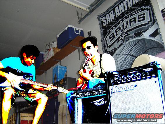 Two musicians play electric guitars in a garage with a San Antonio Spurs banner