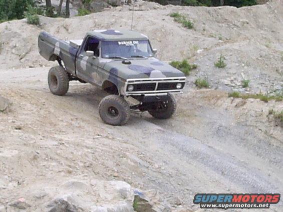 1977 Ford F-150 pickup truck with camouflage paint, lifted suspension, and large tires driving on a dirt hill