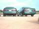1995 Ford Taurus - Two green Ford Taurus SHO sedans are parked on a dirt road, showing their rear ends under a bright sky.