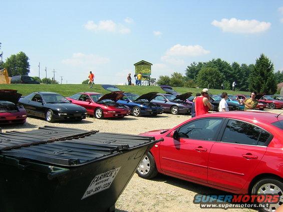 Several cars parked outside with hoods up, people milling around, and a dumpster in the foreground