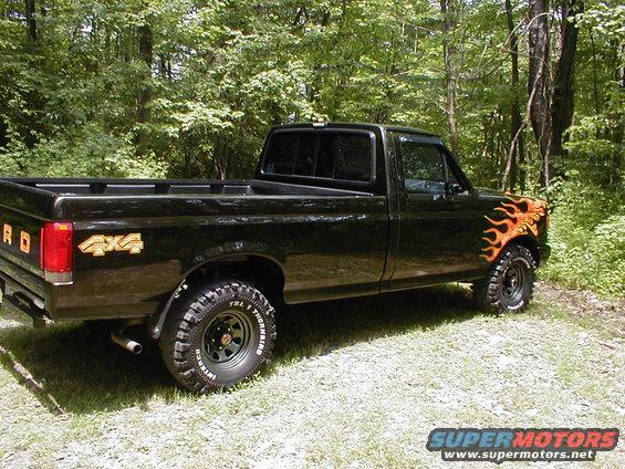 Black 1991 Ford F-150 pickup truck parked in a wooded area, showing flame decals on the front fender and 4x4 badging on the rear quarter panel
