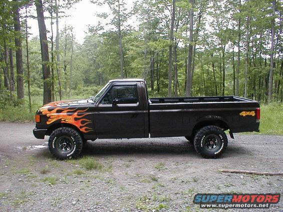 Black Ford F-150 pickup truck with orange and yellow flame decals on the front fender and hood, lifted with off-road tires, parked on a gravel road
