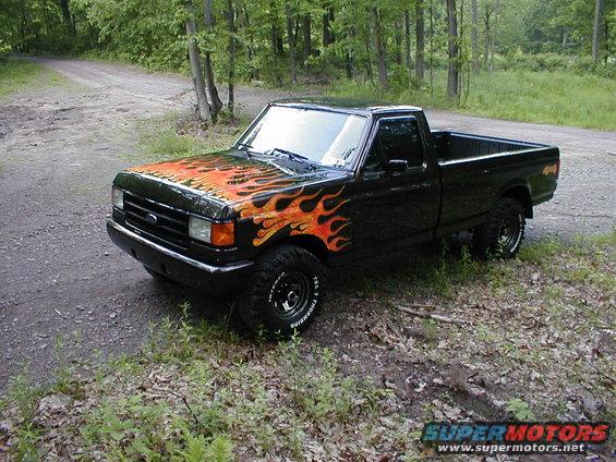 Black pickup truck with flame decals on hood and fender, parked on a dirt road in the woods