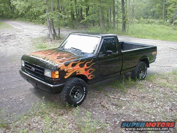 Black 1991 Ford F-150 pickup truck with flame decals on the hood and fender, parked on a dirt road