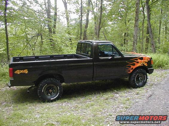 Black 1991 Ford F-150 pickup with flame decals on the fender and 4x4 decal on the rear quarter panel, parked on a dirt road in a wooded area