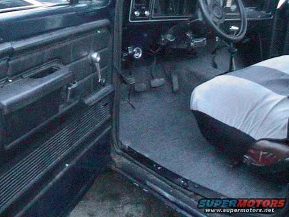 Truck interior showing door panel, steering wheel, pedals, and seat with cover. Appears to be from the mid-1970s