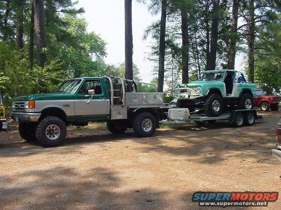 Green Ford Bronco on a trailer, towed by a green and silver Ford F-Series flatbed truck. Another red vehicle is parked behind