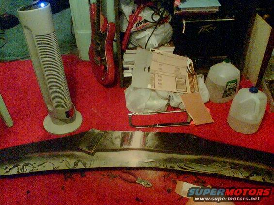 Car bumper being worked on, with tools, sanding paper, and jugs of liquid nearby. Guitars and a fan are in the background