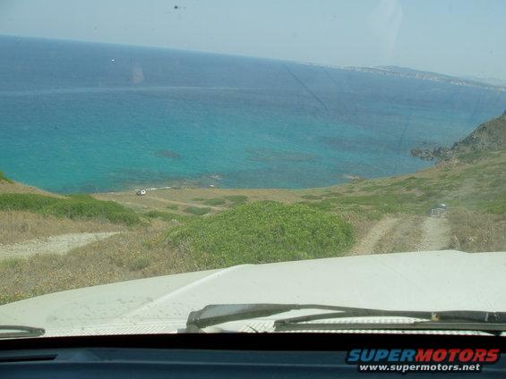 View from inside a vehicle looking out at a coastal road with the ocean on the left. Two other vehicles are visible on the road ahead and near the water