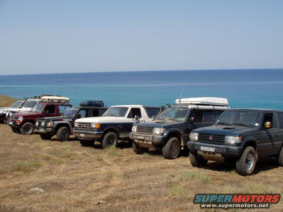 Several off-road SUVs parked in a line on a dry grassy hill with the blue ocean and sky in the background