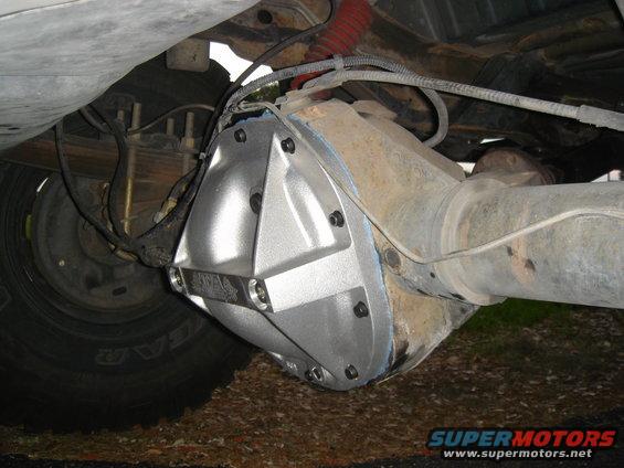 Aftermarket differential cover bolted onto a rusty rear axle housing. Leaf springs and part of a tire are visible