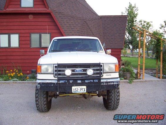 Front view of a white Ford Bronco with a bull bar and off-road tires parked in front of a red house