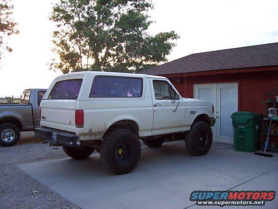 White lifted 1992 Ford Bronco parked next to a red garage, with a silver pickup truck to its left